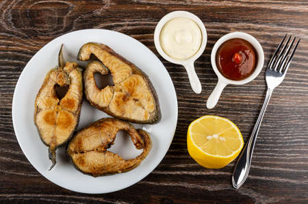 Fried steak from salmon in white plate, sauce boats with mayonnaise and ketchup, half of lemon, fork...