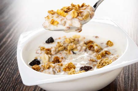 Metallic spoon with cereal breakfast above bowl with granola and yogurt on dark wooden table