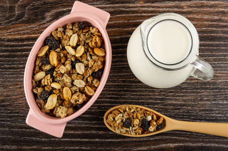Cereal breakfast with peanut and raisin in pink bowl, transparent pitcher with yogurt, plastic spoon with granola on dark wooden table. Top viewの写真素材