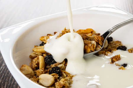 Pouring yogurt in white bowl with cereal breakfast with peanut, raisin on dark wooden tableの写真素材