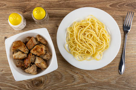 Pepper and salt shakers, glass white bowl with pieces fried pork meat, plate with spaghetti, fork on wooden table. Top viewの写真素材