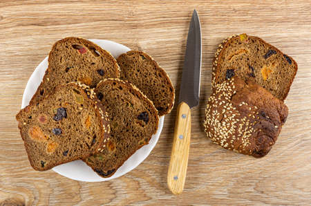 Few slices of bread with fruits and seed in white plate, kitchen knife, bread on wooden table. Top...
