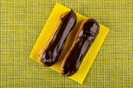 Two eclairs with chocolate glaze on yellow paper napkin on green mat. Top view