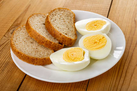 White plate with halves of boiled eggs and slices of bread on wooden table