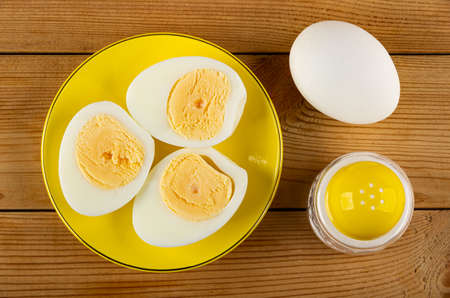 Glass yellow saucer with halves of boiled eggs, unpeeled egg and salt shaker on wooden table. Top...