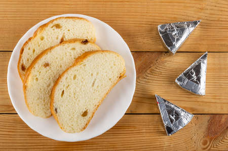 Slices of bread with raisin in white plate, slices of creamy cheese in foil on wooden table. Top viewの写真素材