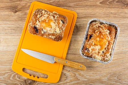 Baked muffin with raisin, kitchen knife on orange cutting board, muffin in foil mold on wooden...