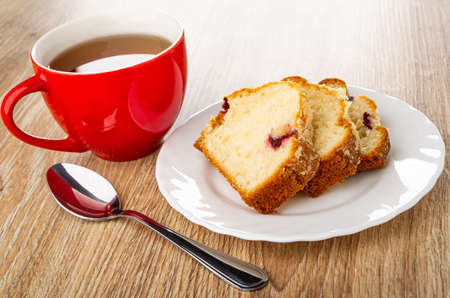 Red cup with tea, muffin with raisin in white plate, teaspoon on wooden table