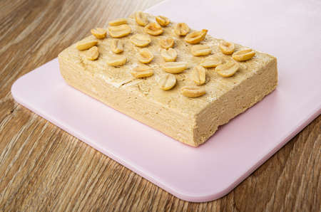 Fried peanut on surface piece of peanut halva on pink plastic cutting board on wooden table