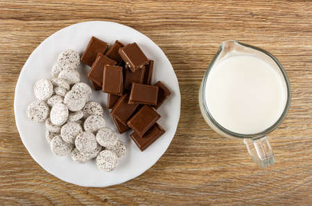 Candy from powdered milk and chocolate in shape tablets, pieces of dairy chocolate in white plate, pitcher with milk on wooden table. Top viewの写真素材