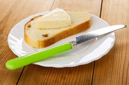Slice of creamy cheese on slice of bread with raisin, table knife in white plate on wooden tableの写真素材