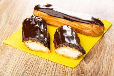 Whole eclair and halves of eclair with chocolate glaze on yellow paper napkin on wooden table