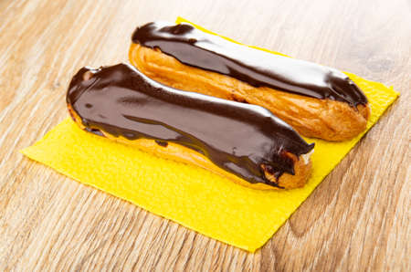 Two eclairs with chocolate glaze on yellow paper napkin on wooden table