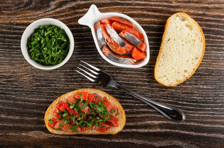 White bowl with chopped parsley, canned salted herring - imitation salmon in bowl in shape fish, slices of wheat bread, fork, sandwich with herring and parsley on wooden table. Top viewの写真素材