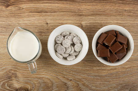 Pitcher with milk, white bowl with candy from powdered milk and chocolate in shape tablets, bowl with dairy chocolate on wooden table. Top view