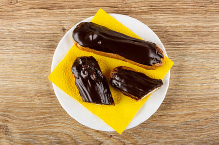 Whole eclair and halves of eclair with chocolate glaze on yellow paper napkin in white plate on...