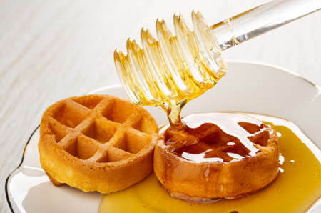 Pouring honey with plastic honey dipper on soft round waffle in white saucer on wooden table