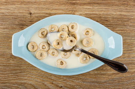 Metallic spoon in blue oval bowl with yogurt and oat rings on wooden table. Top view