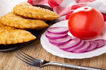 Checkered napkin, fried cheburek in oval brown dish, white plate with tomato and slices of red onion, fork on wooden tableの写真素材