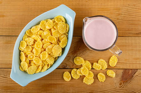 Corn flakes in blue oval bowl, pitcher with pink fruit yogurt, scattered cornflakes on wooden table....