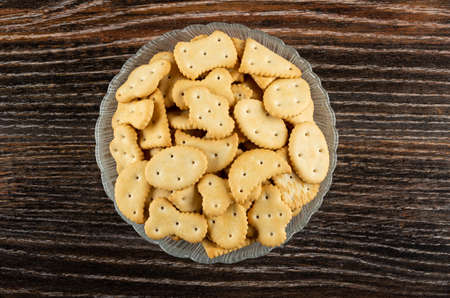 Small crackers different shape in glass bowl on dark wooden table. Top view