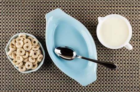 Small oat rings in bowl, metallic spoon in blue oval bowl, jug with yogurt on mat. Top viewの写真素材
