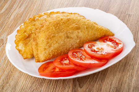 Two fried cheburek, slices of tomato in glass white dish on wooden tableの写真素材