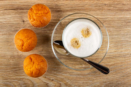 Three orange muffins, transparent glass with latte-macchiato, teaspoon on saucer on wooden table....