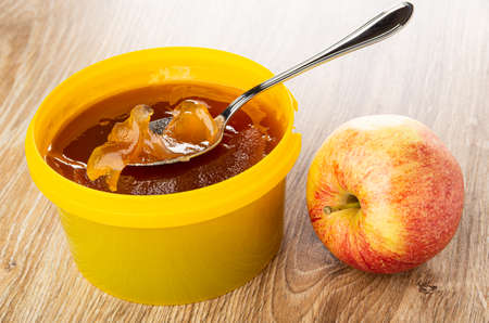 Spoon in apple marmalade in yellow plastic jar, red striped apple on brown wooden table
