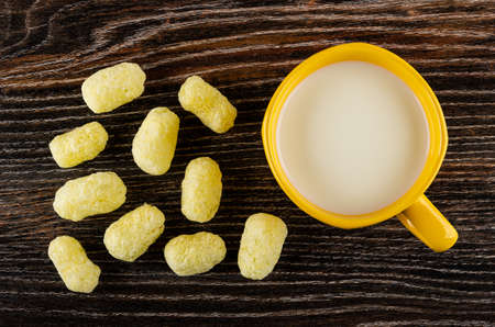 Scattered sweet corn sticks, yellow cup with milk on dark wooden table. Top view