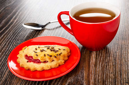 Teaspoon, shortbread cookie with raspberry jam and linseeds in glass saucer, red cup with tea on dark wooden tableの写真素材