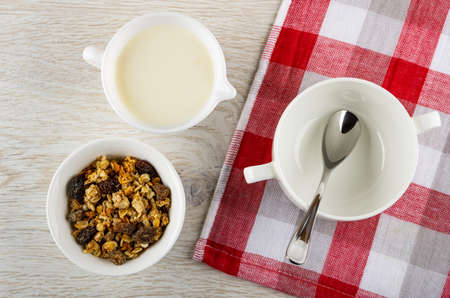 White bowl with baked muesli, pitcher with yogurt, teaspoon in bowl on checkered napkin on wooden table. Top viewの写真素材