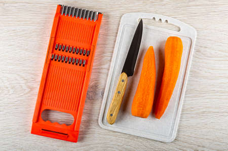 Orange color grater, kitchen knife and two peeled carrots on plastic cutting board on wooden table. Top viewの写真素材