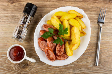 Bottle with condiment, sauce boat with ketchup, glass white dish with slices of fried potatoes, leaves parsley, pieces of sausage, fork on wooden table. Top viewの写真素材