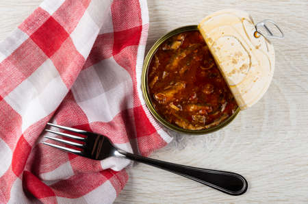 Checkered napkin, fork, opened metallic jar with sprat in tomato sauce on wooden table. Top viewの写真素材
