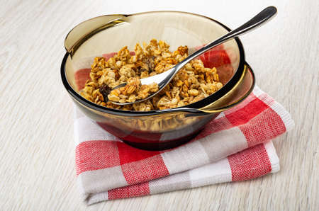 Teaspoon in transparent brown bowl with baked muesli on folded napkin on wooden table
