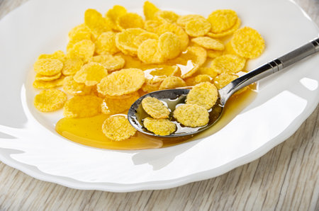 Close up of teaspoon in white plate with corn flakes and liquid honey on wooden table