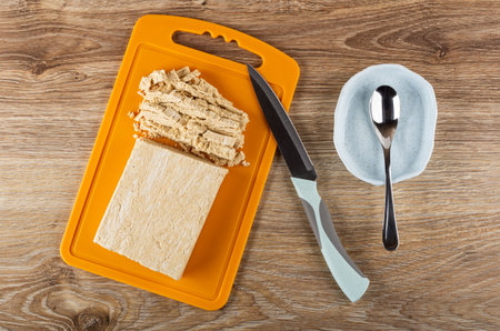 Pieces of cutted peanut halva on orange plastic cutting board, kitchen knife, teaspoon in light-blue...