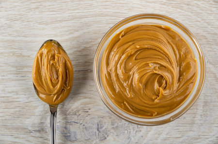 Metallic spoon with peanut butter, transparent glass bowl with peanut butter on wooden table. Top...