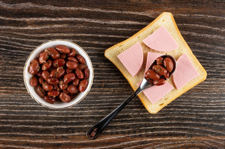White bowl with lobio, teaspoon with red bean on sandwich with sausage on dark wooden table. Top viewの写真素材