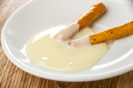 Two breadsticks poured condensed milk in white saucer on brown wooden table