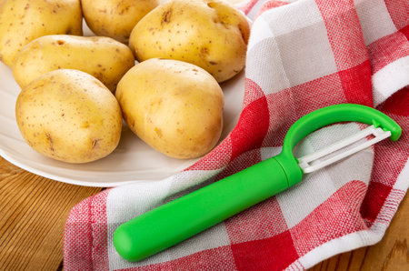 Raw potatoes with peel in white glass dish and peeler on checkered napkin on wooden tableの写真素材