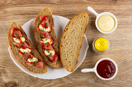 Sandwiches from rye bread with sausage, mayonnaise and ketchup, slice of bread in white dish, sauce boats with sauce, pepper shaker on wooden table. Top viewの写真素材