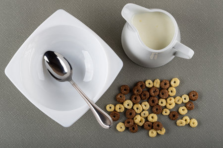 Spoon in white glass bowl, pitcher with yogurt, scattered toasted cereal breakfasts with chocolate and caramel on gray table. Top viewの写真素材