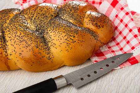 Wicker bun with poppy seeds on checkered napkin, kitchen knife on wooden table