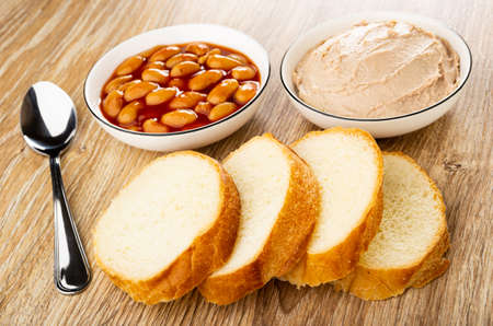 Teaspoon, meat pate in glass bowl, canned beans in bowl, slices of wheat bread on wooden table