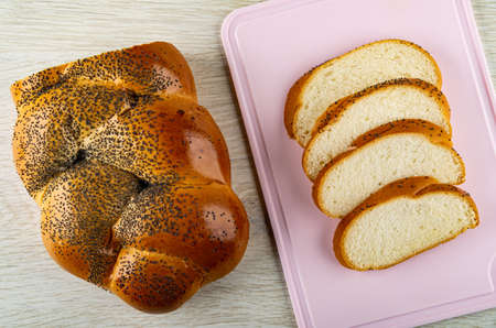 Piece of wicker bun with poppy seeds, slices of bun on pink plastic cutting board on wooden table....