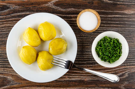 Baked potatoes in white plate, fork, salt in bamboo bowl, glass bowl with chopped parsley on dark wooden table. Top viewの写真素材