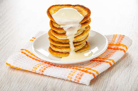 Stack of small pancakes poured condensed milk in white saucer on folded checkered napkin on wooden...