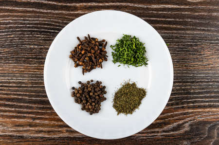 Small heaps of clove spice, peppercorn, dried parsley and dried dill in white plate on wooden table. Top viewの写真素材